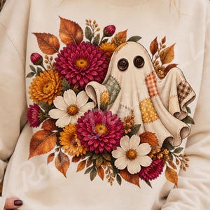 Peut inclure: Un sweat-shirt beige avec une impression graphique représentant un fantôme en tissu patchwork doté de deux boutons marron foncé en guise d'yeux. Le fantôme est entouré d'un arrangement floral automnal composé de dahlias rouge foncé, de chrysanthèmes jaunes et de marguerites blanches, agrémenté de feuilles d'automne brunes et orange.