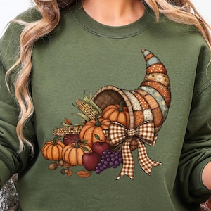 May include: Olive green sweatshirt featuring a colorful cornucopia design. The cornucopia is filled with pumpkins, apples, grapes, and corn. A brown and white checkered bow adorns the front of the cornucopia. The horn is made of patchwork fabric.