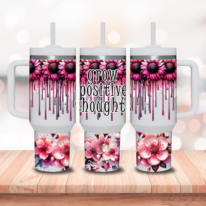 May include: Three white stainless steel tumblers with pink floral designs and dripping pink paint. The middle tumbler has the text "grow positive thought" in black.