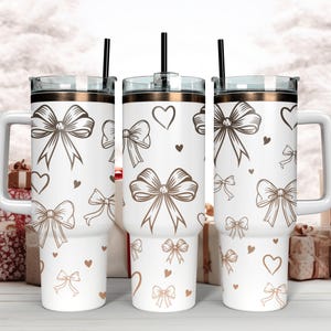 Puede incluir: Vaso blanco con un patrón de lazo y corazón marrón. El vaso tiene un asa, una pajita negra y un borde marrón. El diseño presenta varios estilos de lazos y pequeños corazones.