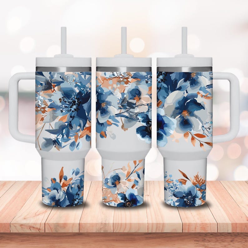 May include: Three white travel mugs with blue and orange floral designs. Each mug has a lid and a straw. The mugs are on a wooden surface.