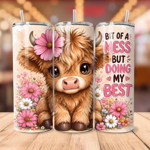 May include: A cylindrical tumbler with a straw featuring a fluffy brown highland cow illustration, pink and white flowers, and the text "BIT OF A MESS BUT DOING MY BEST" in pink lettering against a light beige background with soft watercolor accents.