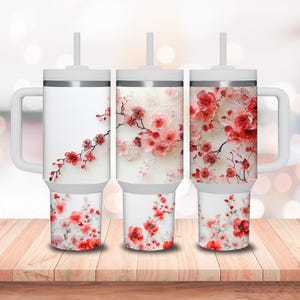 May include: Three white stainless steel tumblers with a floral design. Each tumbler has a lid and a straw. The tumblers are on a wooden surface.