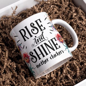 May include: White ceramic coffee mug with a black and white graphic that says "Rise and Shine...mother cluckers." The graphic is surrounded by colourful floral designs.