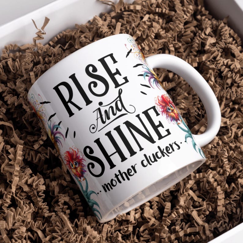 May include: White ceramic coffee mug with a black and white graphic that says "Rise and Shine...mother cluckers." The graphic is surrounded by colourful floral designs.