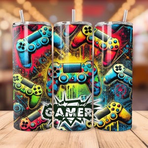 May include: A colorful, abstract design with multiple video game controllers on a black background. The word "GAMER" is in a green and black starburst shape. This design is printed on a stainless steel tumbler.