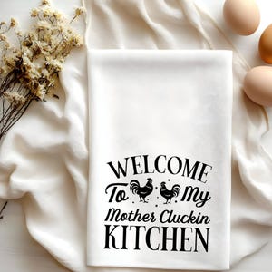 Puede incluir: Toalla de cocina blanca con texto negro que dice "Bienvenido a mi cocina de madre Cluckin" con dos siluetas de gallos negros.