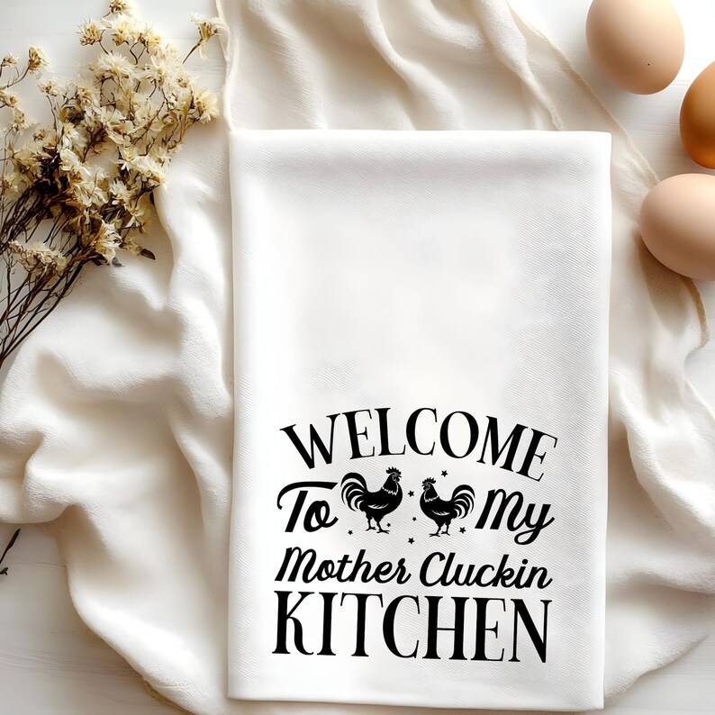 Puede incluir: Toalla de cocina blanca con texto negro que dice "Bienvenido a mi cocina de madre Cluckin" con dos siluetas de gallos negros.