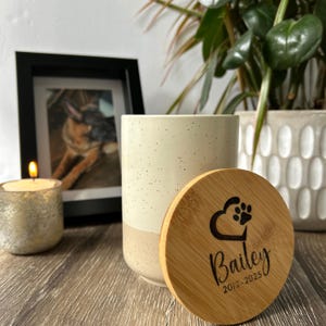 May include: A white ceramic jar with a speckled finish and a wooden lid. The lid has a paw print heart design and the text "Bailey 2012-2025".