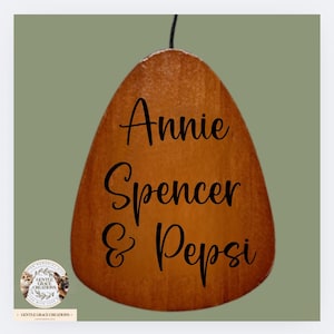 May include: A wooden memorial ornament with the names "Annie Spencer & Pepsi" in black script. The ornament is teardrop-shaped and hangs from a black string. The background is a muted green.