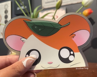 May include: A sticker of a cartoon hamster with orange fur, a white face, and big black eyes. The hamster is wearing a green leaf on its head. The sticker is on a wooden surface.