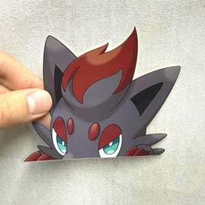 Puede incluir: Una pegatina con un personaje de dibujos animados con aspecto de zorro, con pelaje gris, detalles rojos y ojos turquesa. El personaje tiene orejas puntiagudas y un mechón de pelo rojo. La pegatina está sujeta por una mano.