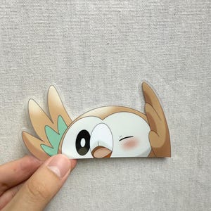 May include: A die-cut vinyl sticker featuring a cartoon owl character with tan and mint green plumage. The character has one large black eye, a small beak, and a winking eye with a soft pink blush on its cheek, depicted in a playful pose.