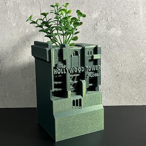 May include: A green 3D printed planter shaped like the Hollywood Tower Hotel. The planter has the words "The Hollywood Tower Hotel" on the front. A small green plant is growing in the planter.