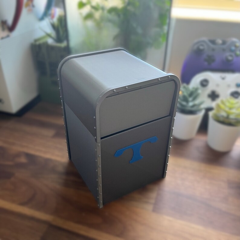 Puede incluir: Una caja gris con bordes met&aacute;licos y una letra "T" azul en la parte delantera. La caja tiene una tapa con bisagras y parece ser un contenedor de almacenamiento.