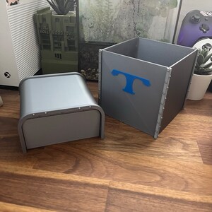 Puede incluir: Dos cajas de almacenamiento grises con un acabado met&aacute;lico. La caja m&aacute;s grande tiene una letra "T" azul en la parte delantera. La caja m&aacute;s peque&ntilde;a tiene una tapa con bisagras.