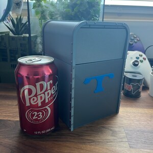 Puede incluir: Una caja de metal gris con una "T" azul en la parte delantera. La caja est&aacute; sobre una superficie de madera junto a una lata de Dr Pepper.