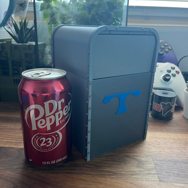 Puede incluir: Una caja de metal gris con una "T" azul en la parte delantera. La caja est&aacute; sobre una superficie de madera junto a una lata de Dr Pepper.