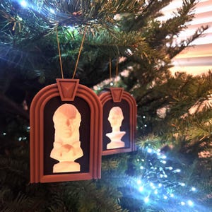 May include: Two brown framed Christmas ornaments with white 3D busts inside. The ornaments are hanging on a Christmas tree.