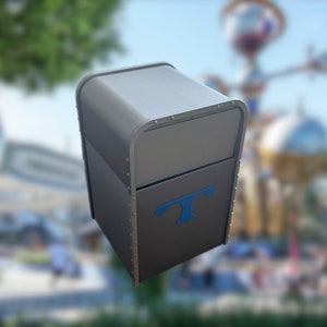 Puede incluir: Un contenedor de basura de metal gris con una "T" azul en la parte delantera. El contenedor de basura tiene una tapa con bisagras y una superficie texturizada.