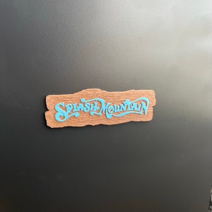 May include: A wooden sign with a blue "Splash Mountain" logo. The sign is a light brown color and has a rough, textured surface.