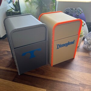 Puede incluir: Dos cajas decorativas con acabado gris y beige. Las cajas tienen una tapa con bisagras y una peque&ntilde;a manija en la parte delantera. La caja gris tiene una T azul en la parte delantera. La caja beige tiene la palabra "Disneyland" en azul en la parte delantera.