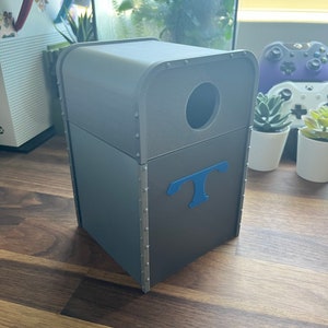 Puede incluir: Un cubo de basura rectangular gris con una abertura redonda en la parte superior y una letra "T" azul en la parte delantera. El cubo de basura est&aacute; hecho de un material texturizado y tiene un borde met&aacute;lico alrededor de la parte superior.