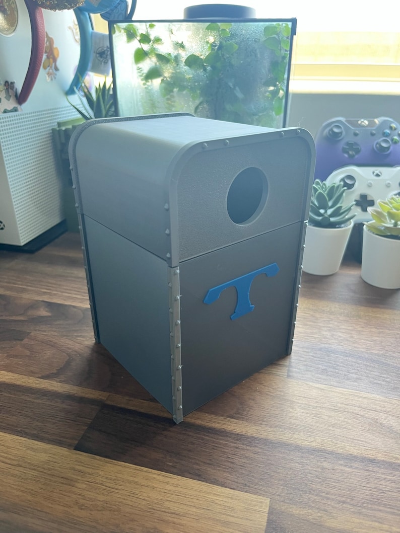 Puede incluir: Un cubo de basura rectangular gris con una abertura redonda en la parte superior y una letra "T" azul en la parte delantera. El cubo de basura est&aacute; hecho de un material texturizado y tiene un borde met&aacute;lico alrededor de la parte superior.