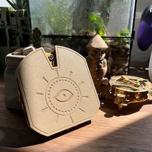 May include: A beige, 3D printed, decorative box with a sun and eye design. The box is closed and has a small, black, metal latch.