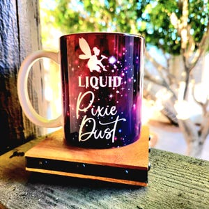 May include: A white ceramic mug with a handle. The mug has a galaxy-themed design with pink, purple, and blue colors. The text "Liquid Pixie Dust" is printed on the mug in white.