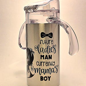 May include: Stainless steel sippy cup with a clear plastic lid and handles. The cup features the text "future ladies man currently mama's boy" in black and blue script, along with a bow tie graphic and a silhouette of a boy.