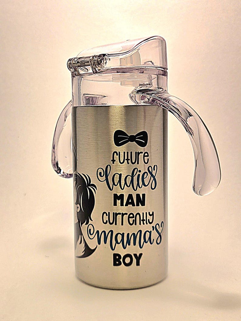 May include: Stainless steel sippy cup with a clear plastic lid and handles. The cup features the text "future ladies man currently mama's boy" in black and blue script, along with a bow tie graphic and a silhouette of a boy.