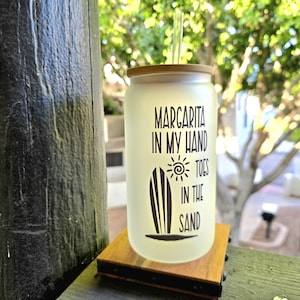 May include: A frosted glass tumbler with a bamboo lid and a clear straw. The tumbler has the words "Margarita in my hand, toes in the sand" printed in black, along with a surfboard and sun graphic. The tumbler sits on a wooden coaster.