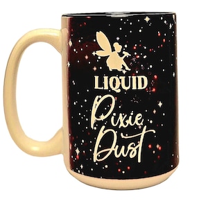 May include: A ceramic mug with a cream-colored handle and rim. The mug features a black background with white stars and a fairy illustration. The words "LIQUID Pixie Dust" are written in white script.