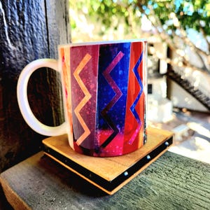 May include: A colorful ceramic mug with a geometric pattern in shades of red, blue, yellow, and purple. The mug is sitting on a wooden coaster with black accents.
