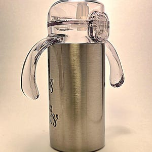 May include: Stainless steel sippy cup with a clear plastic lid, handles, and straw. The cup has a brushed metal finish and features black cursive text. The lid has a flip-top closure for easy access.