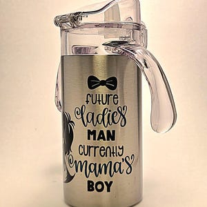 May include: Stainless steel sippy cup with a clear plastic lid and handle. The cup features the text "Future Ladies Man Currently Mama's Boy" in black script and a black bow tie graphic.