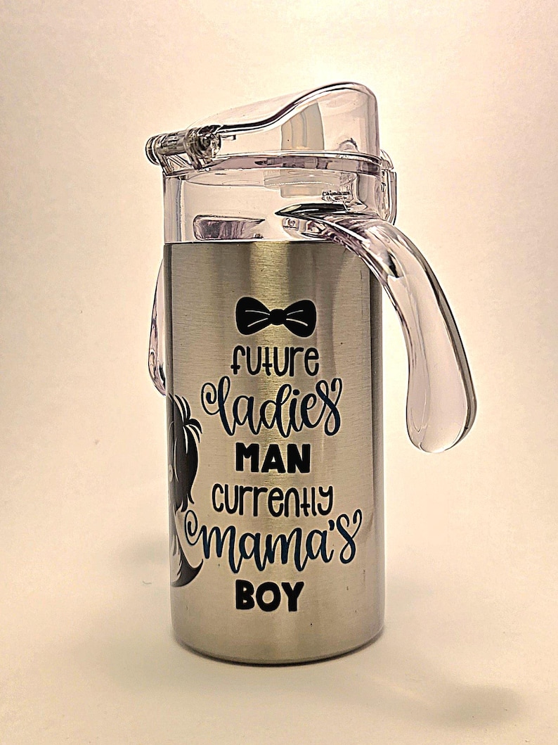 May include: Stainless steel sippy cup with a clear plastic lid and handle. The cup features the text "Future Ladies Man Currently Mama's Boy" in black script and a black bow tie graphic.