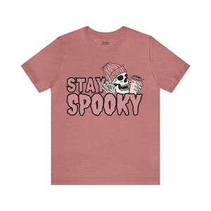 May include: A pink heather t-shirt featuring a graphic of a skeleton wearing a pink beanie and holding a pink cup. The text "STAY SPOOKY" is printed in pink letters on the shirt.