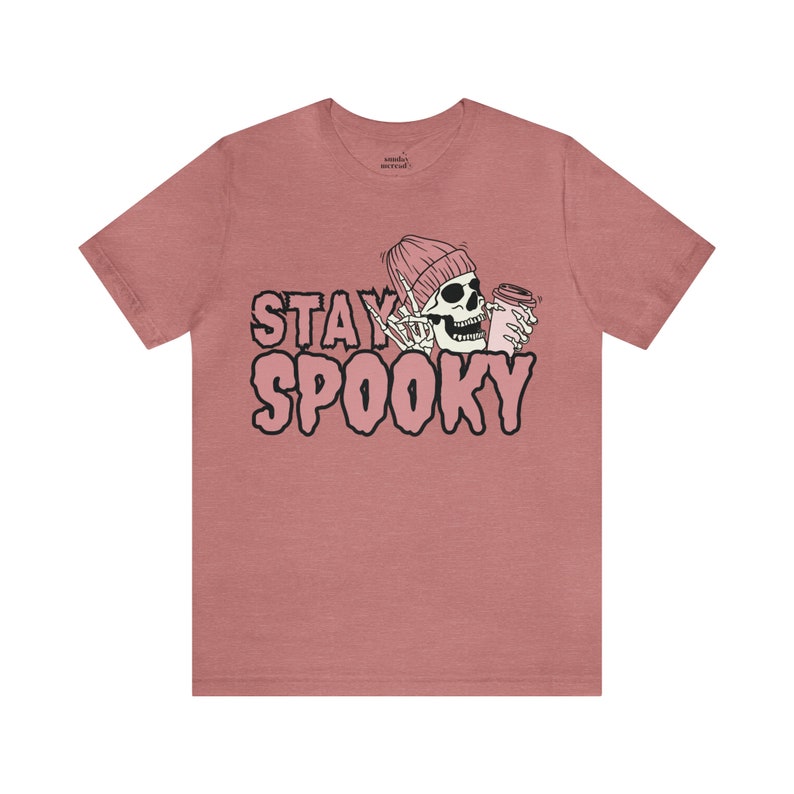 May include: A pink heather t-shirt featuring a graphic of a skeleton wearing a pink beanie and holding a pink cup. The text "STAY SPOOKY" is printed in pink letters on the shirt.
