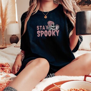 May include: A black t-shirt with the text "STAY SPOOKY" in pink lettering. A pink skull wearing a pink beanie is holding a mug of coffee.