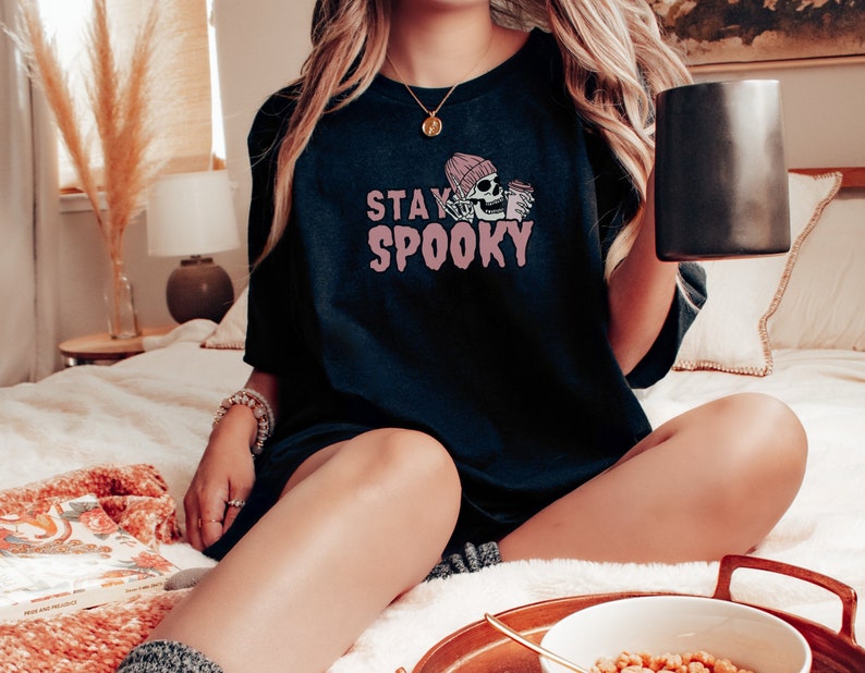 May include: A black t-shirt with the text "STAY SPOOKY" in pink lettering. A pink skull wearing a pink beanie is holding a mug of coffee.