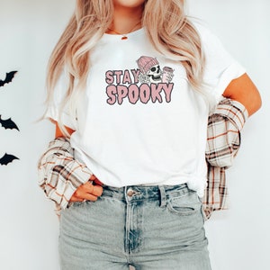 May include: White t-shirt featuring a pink graphic of a skeleton wearing a pink beanie and holding a coffee cup. The text "STAY SPOOKY" is written in pink letters.