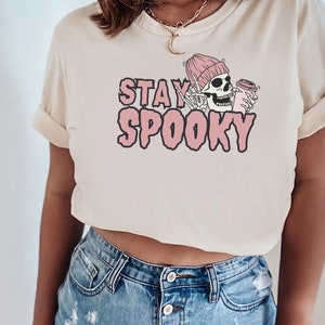 May include: A cropped, cream-coloured t-shirt with a pink graphic of a skeleton wearing a pink beanie and holding a coffee cup. The text "STAY SPOOKY" is written in pink letters.