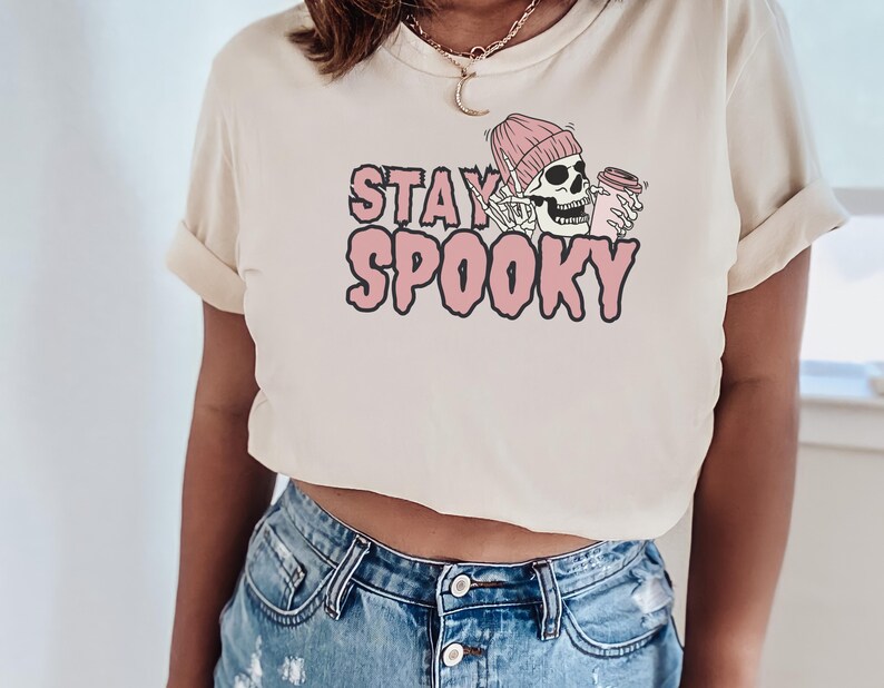 May include: A cropped, cream-coloured t-shirt with a pink graphic of a skeleton wearing a pink beanie and holding a coffee cup. The text "STAY SPOOKY" is written in pink letters.