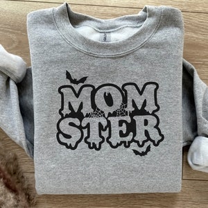 Peut inclure: Sweat-shirt gris avec un motif noir qui dit "MOMSTER" avec des lettres qui coulent et des chauves-souris.
