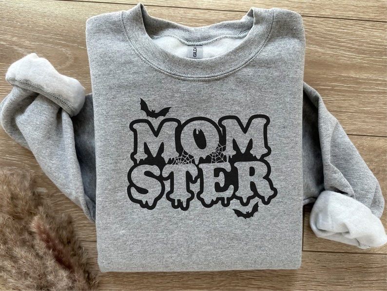 Peut inclure: Sweat-shirt gris avec un motif noir qui dit "MOMSTER" avec des lettres qui coulent et des chauves-souris.