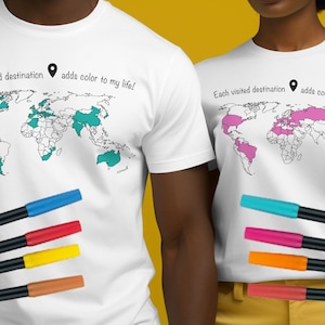 May include: White t-shirts with a world map design, featuring countries colored in teal and pink. The text "Each visited destination adds color to my life!" is printed above the map. Several colored markers are in the foreground.