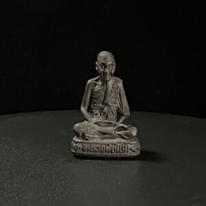 Puede incluir: Una pequeña estatua de un monje budista sentado, de color plateado. El monje lleva una túnica y tiene una expresión serena en su rostro. La estatua está sentada sobre una base con escritura tailandesa.