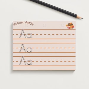 May include: A white card with the text "Autumn ABC's" at the top. The card has three lines with the letter "Aa" written on each line. The lines have brown dashed lines for tracing. There is a small illustration of a stack of books with leaves on top in the upper right corner.
